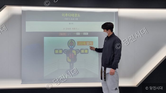 태권도 지도자가 실내에서 겨루기 프로그램 화면을 가리키고 있다.