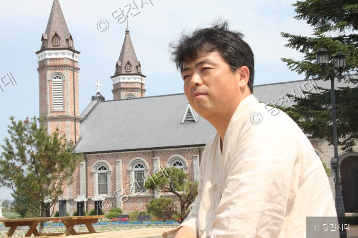 홍광철 합덕성당 주임신부가 성당을 배경으로 포즈를 취하고 있다.