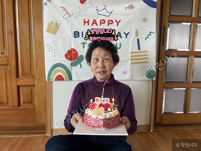 한 어르신이 생일 케이크를 들고 밝은 표정으로 앉아 있다.