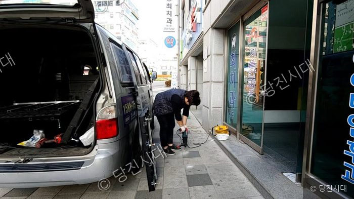작업자가 건물 앞 인도에서 전동공구로 바닥을 뚫고 있다.