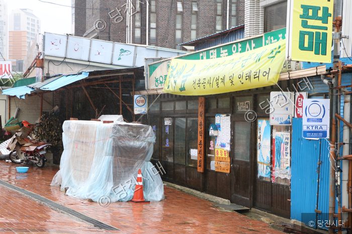 비 오는 날 거리에 상점들과 포장된 짐이 놓여 있다.