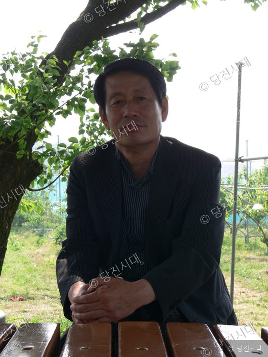 홍천표 전 당진군농업기술센터 소장이 나무 그늘 아래 야외 벤치에 앉아 포즈를 취하고 있다.