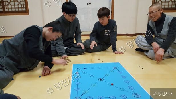 스님과 학생들이 실내에서 윷놀이를 하며 즐거워하고 있다.