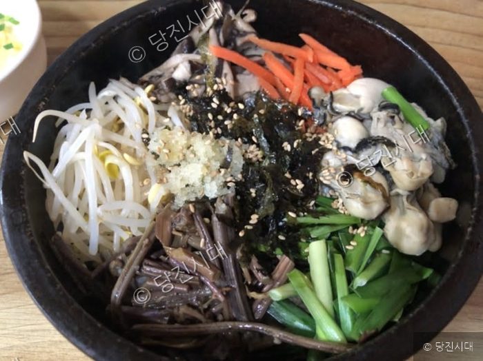 식당에서 신선한 굴과 다양한 나물이 들어간 굴비빔밥이 그릇에 담겨 있다.