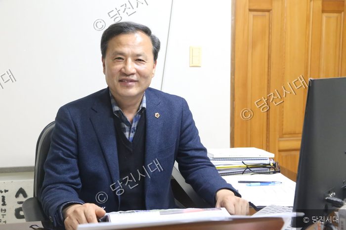 남성 의원이 사무실 책상에 앉아 미소를 짓고 있다.