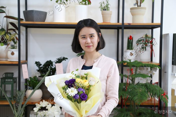 꽃집 대표가 매장에서 꽃다발을 들고 포즈를 취하고 있다.