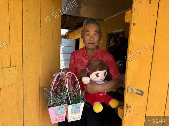 노인이 카네이션 화분과 인형을 들고 집 문 앞에 서 있다.