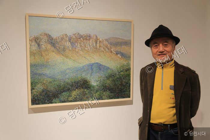 한 남성이 자신이 그린 산 그림 작품 옆에서 포즈를 취하고 있다.