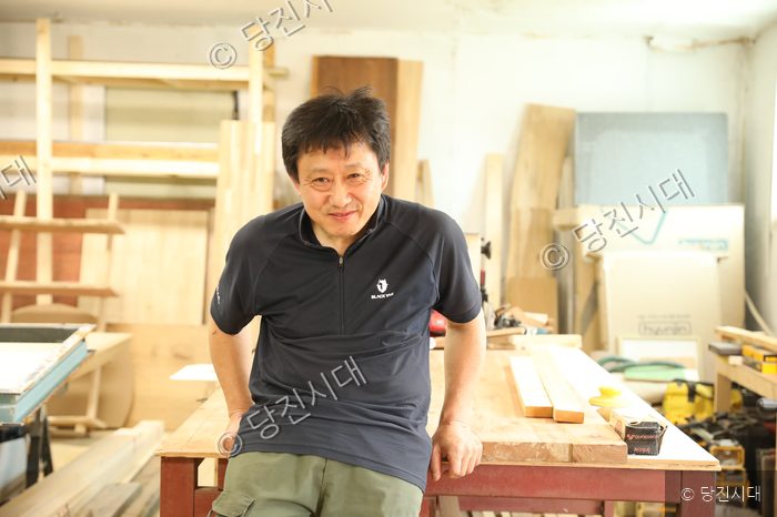 목공소 작업대에 기댄 남성이 미소를 짓고 있다.
