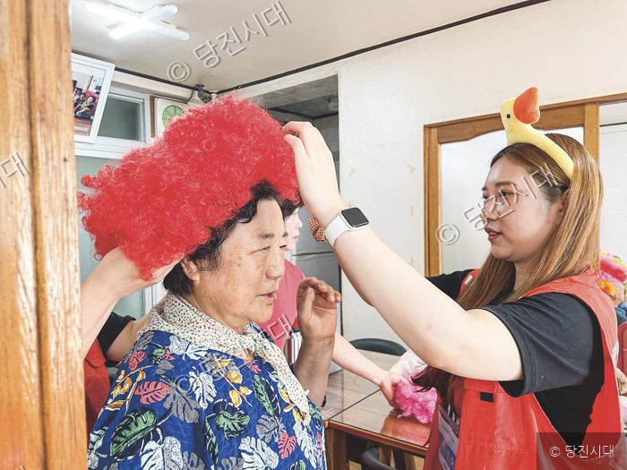 자원봉사자가 어르신에게 빨간 가발을 씌워주고 있다.