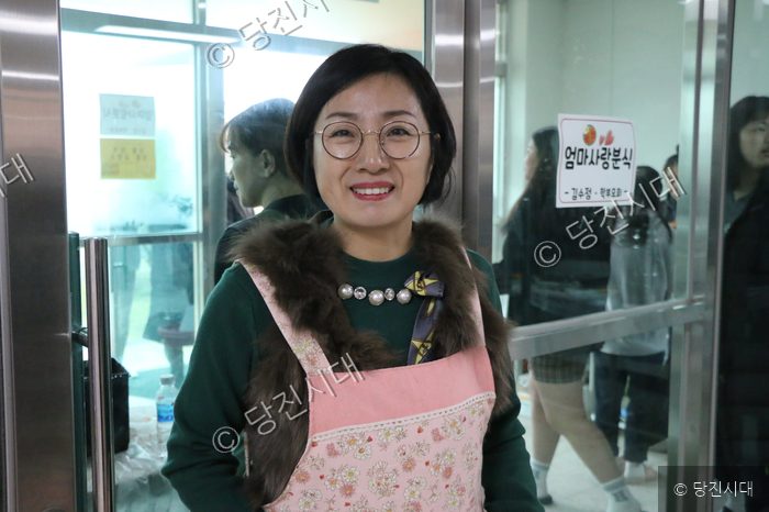 한 여성이 앞치마를 두르고 미소를 지으며 카메라를 바라보고 있다.