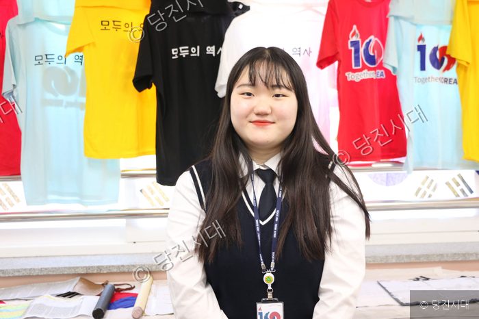 한 여학생이 송악고등학교의 역사 관련 행사 기념 셔츠 앞에서 미소 짓고 있다.