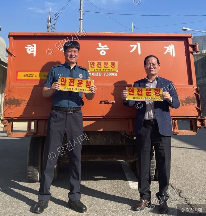 두 남성이 대형 농기계 앞에서 안전운행 표지판을 들고 있다.