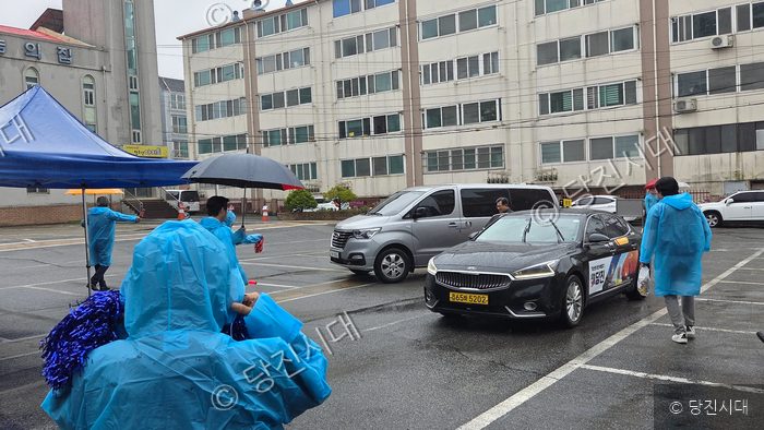 우중 속에서 사람들이 파란 우의를 입고 차량을 맞이하고 있다.