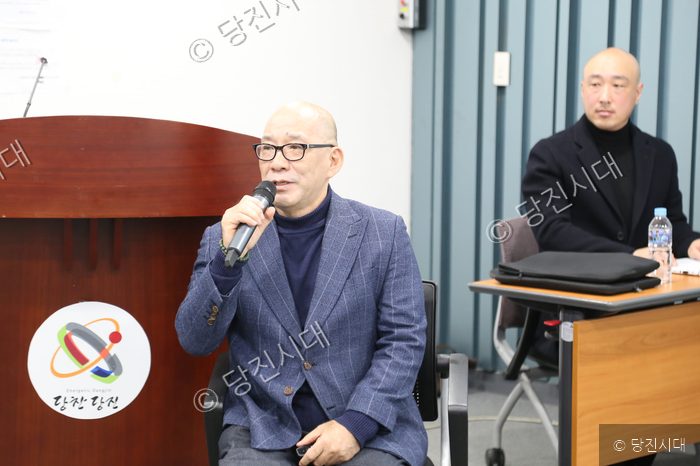 한 남성이 마이크를 들고 발표하고 있고, 옆에는 다른 남성이 앉아 있다.