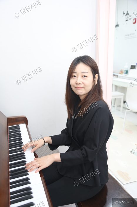 피아노 앞에 앉은 오슬아 원장이 카메라를 바라보며 미소 짓고 있다.