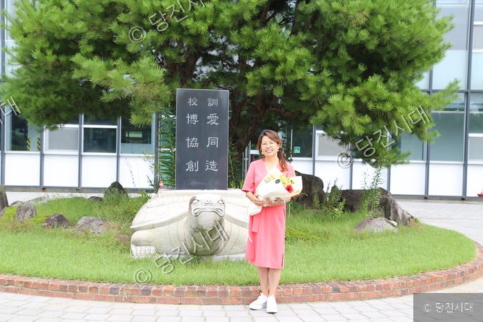 김광자 선생님이 교훈석과 거북상 앞에서 꽃다발을 들고 포즈를 취하고 있다.