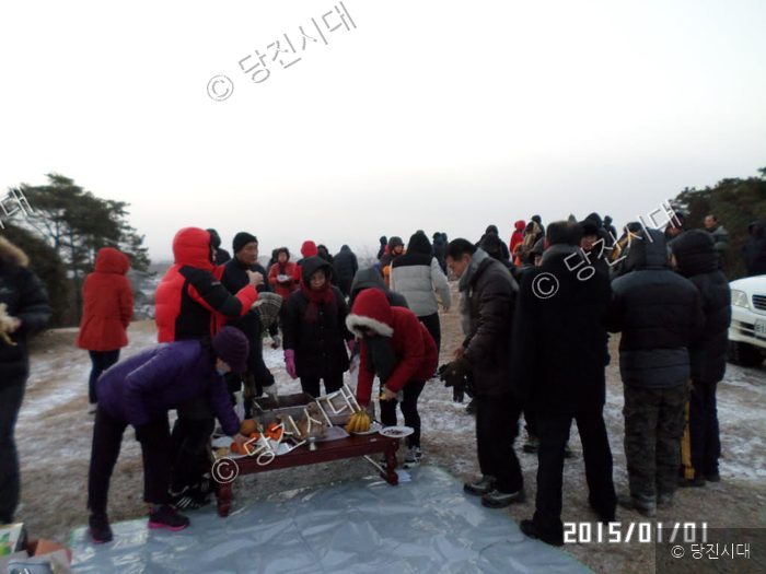 사람들이 야외에 모여 제사상 형태의 차림을 두고 서 있다.