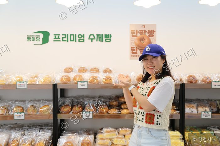 빵집 점장이 진열대 앞에서 빵을 가리키며 미소 짓고 있다.