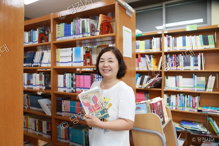 여성이 도서관 서가 앞에서 책을 들고 미소 짓고 있다.