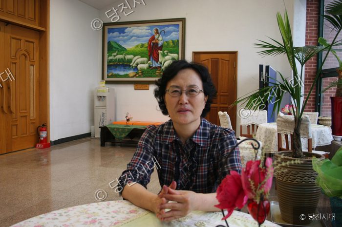 안경을 쓴 여성이 실내 테이블에 앉아 카메라를 바라보고 있다.