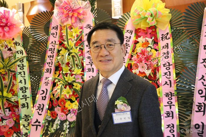 안경을 쓴 남성이 축하 화환을 배경으로 미소를 짓고 있다.