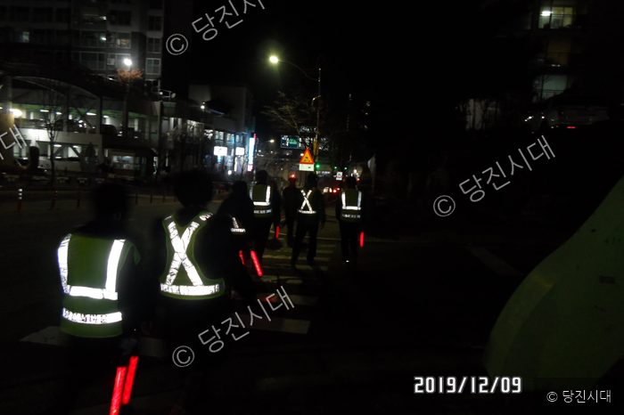야간에 형광 조끼를 입은 사람들이 거리를 걷고 있다.