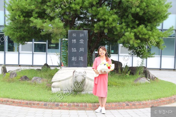 박옥진 씨가 학교 교훈석 앞에서 꽃다발을 들고 포즈를 취하고 있다.
