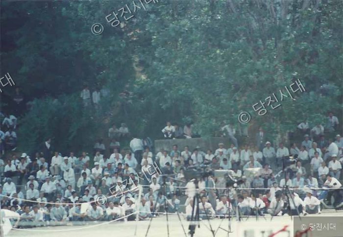 많은 사람들이 야외 유세장에 모여 앉아 있다.
