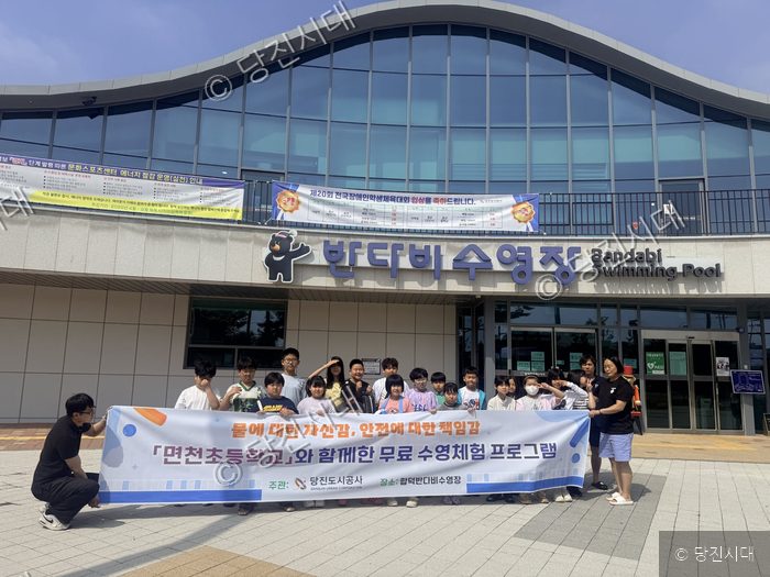 학생들과 인솔자들이 수영장 앞에서 무료 수영체험 기념 현수막을 들고 포즈를 취하고 있다.