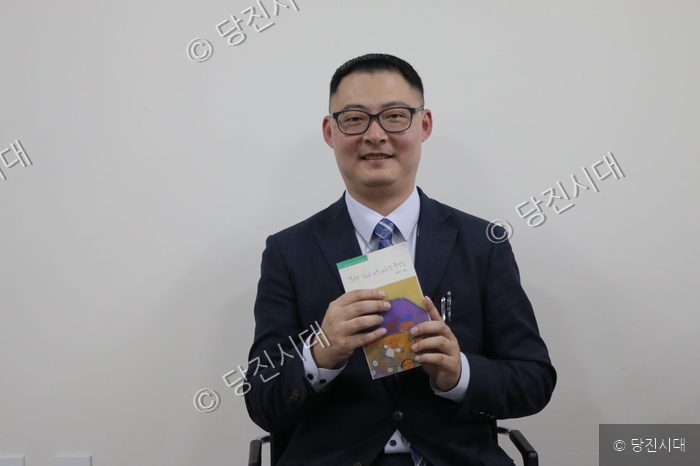 한 남성이 책을 들고 미소 지으며 포즈를 취하고 있다.