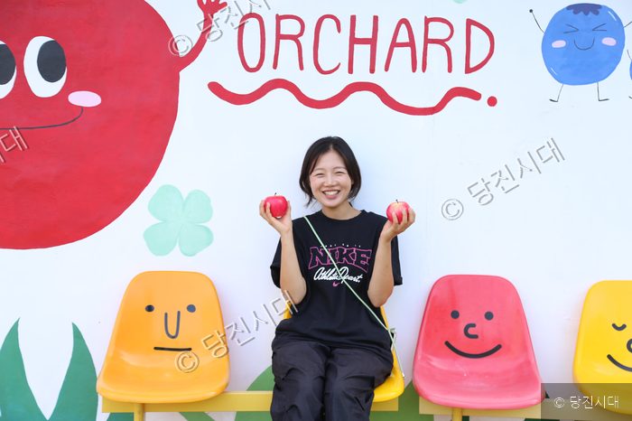 사과 농장 벽화를 배경으로 사과를 든 성은영 대표가 미소 짓고 있다.