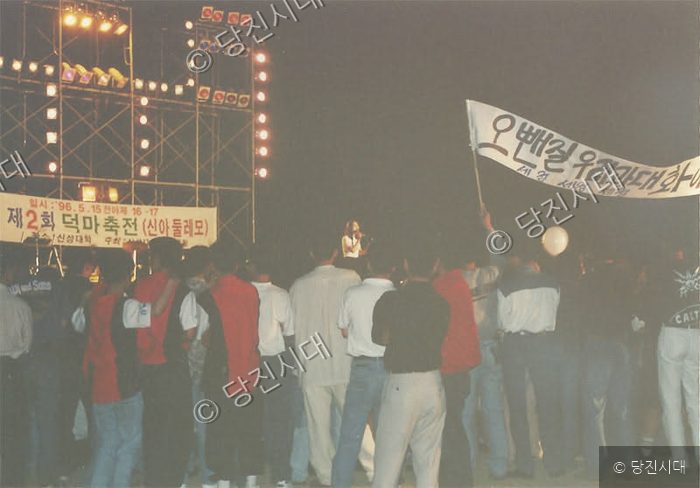 밤거리 야외 무대 앞에서 사람들이 모여 행사를 관람하고 있다.