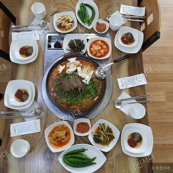 식탁 위에 푸짐하게 차려진 흑염소전골과 다양한 밑반찬들이 놓여 있다.
