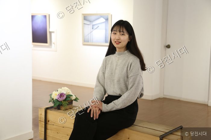 여성 작가가 갤러리 벤치에 앉아 미소 짓고 있다.