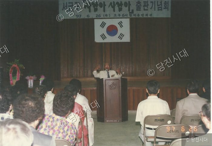 실내 강연장에서 한 인물이 단상에 서서 발표하고 있다.