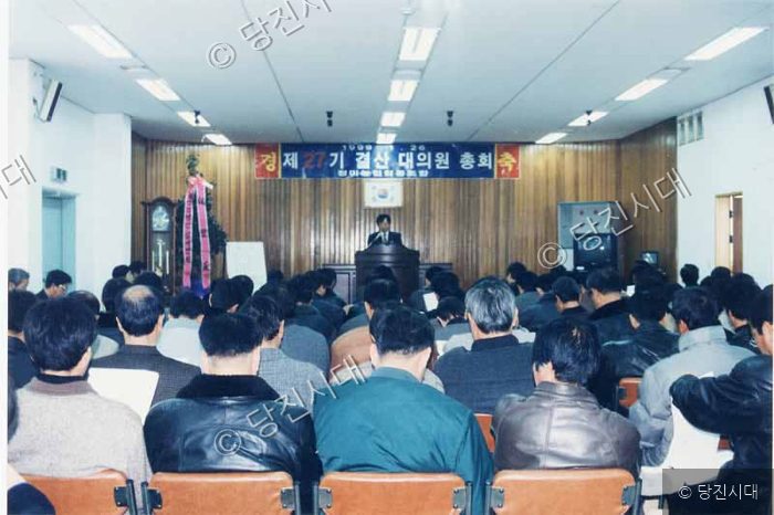 실내 강당에서 많은 사람들이 참석한 가운데 총회가 열리고 있다.