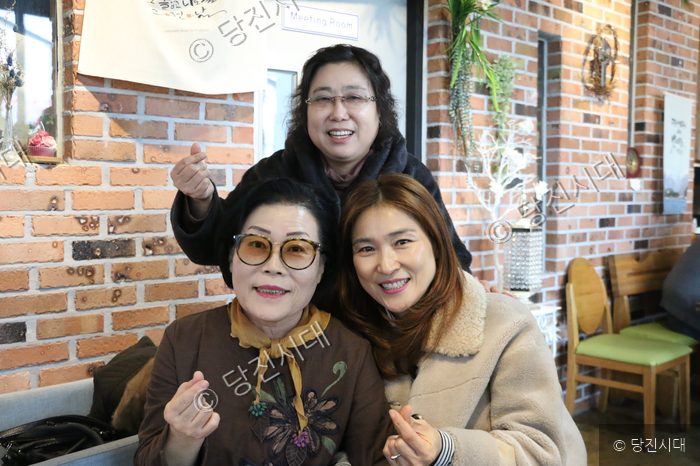 세 명의 여성이 카페에 모여 손가락 하트 포즈를 취하며 미소 짓고 있다.