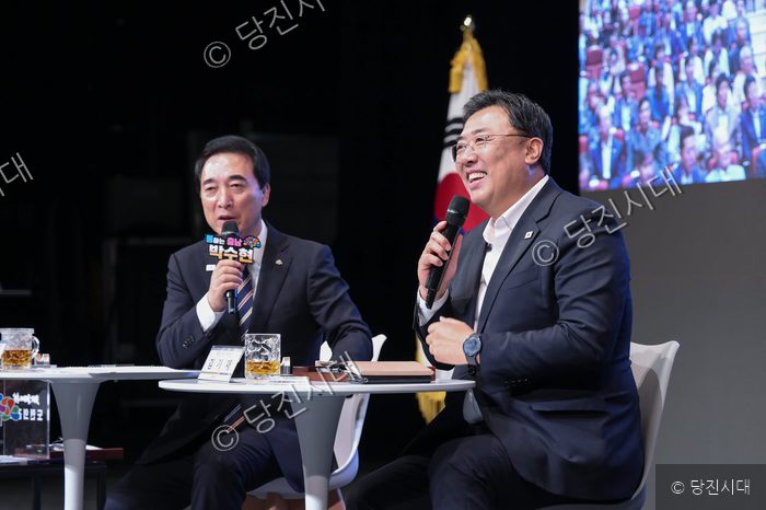 두 남성이 테이블 앞에 앉아 마이크를 잡고 이야기를 나누고 있다.