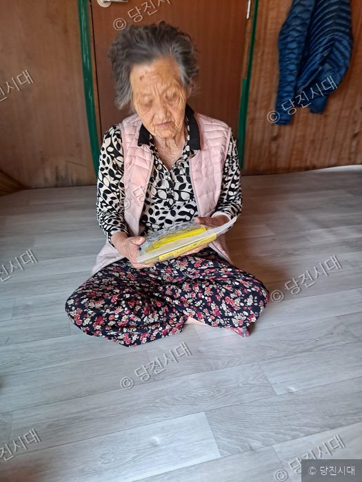 한 노인이 실내에서 바닥에 앉아 물품을 살펴보고 있다.