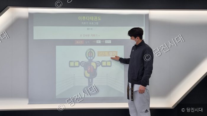 태권도 지도자가 스크린의 겨루기 프로그램을 가리키고 있다.