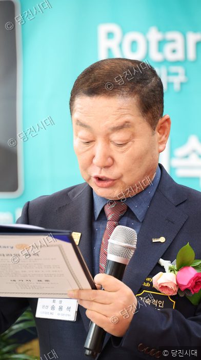 한 남성이 로타리 클럽 행사장에서 마이크를 잡고 문서를 읽고 있다.