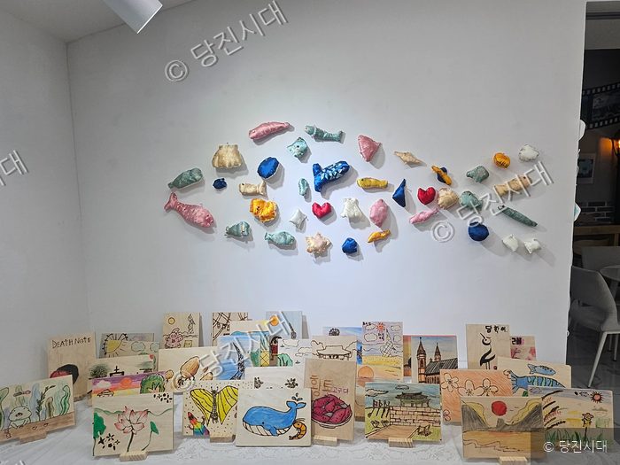 갤러리에 전시된 물고기 모형과 아동화들이 벽면과 탁자에 놓여 있다.