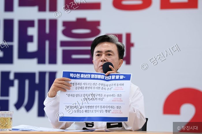 한 남성이 마이크를 잡고 인쇄물을 들어 보이고 있다.
