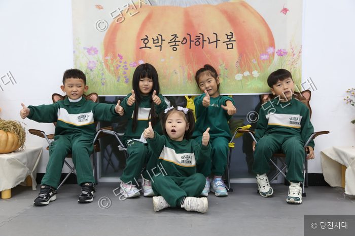어린이들이 초록색 체육복을 입고 호박 그림 앞에서 포즈를 취하고 있다.