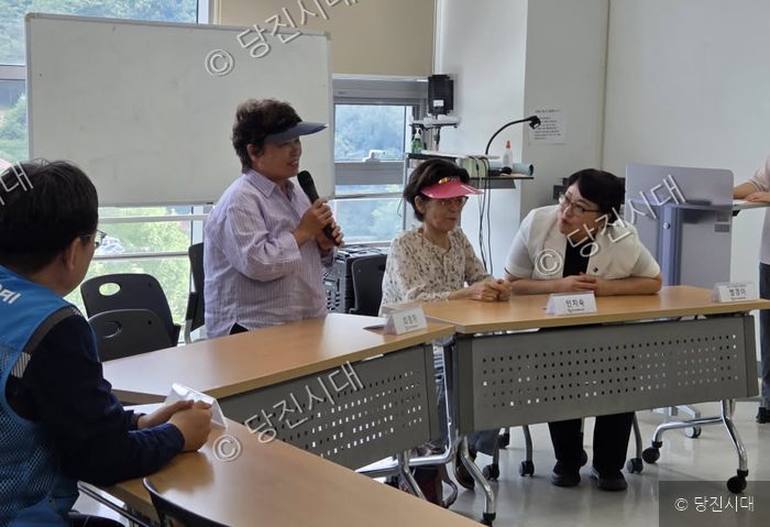 한 여성이 마이크를 잡고 이야기를 하고 있으며 옆에 다른 참석자들이 앉아 있다.