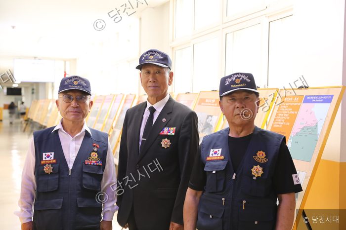 참전유공자 세 사람이 사진전 전시물 앞에서 포즈를 취하고 있다.
